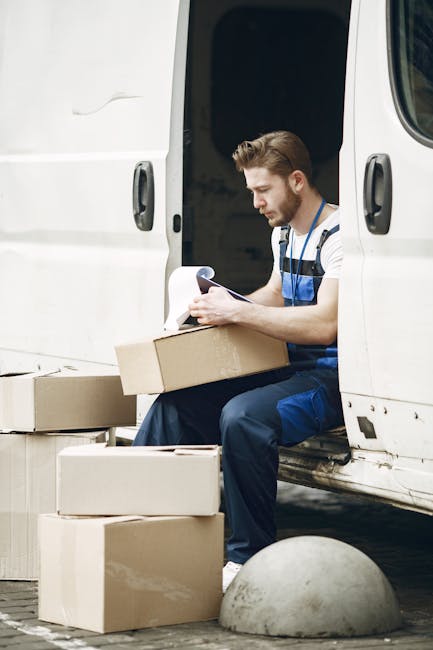 A man wearing a blue jacket and dark work trousers is loading large white cardboard boxes into the back of a white moving van parked on a city street in daylight. The van's rear doors are wide open, r