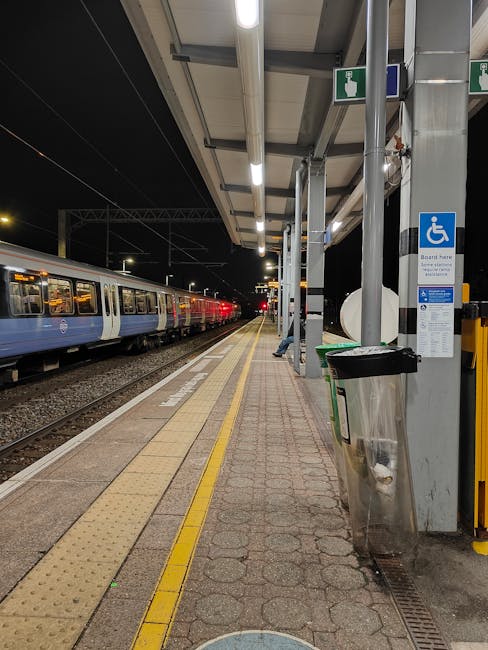Nighttime scene at Lewisham Way train station showing an empty platform with tactile paving and yellow safety lines along the edge. A blue and white train is stationary on the left side, its doors closed. The platform is sheltered by a metal canopy with built-in lighting that illuminates the area. A black and silver trash bin is positioned near a support pillar on the platform, with a blue disabled access sign attached to the pillar. Visible on the platform are a few people, some dressed in high-visibility clothing, engaged in loading or unloading furniture, boxes, and packing materials onto a van parked nearby for a house removal at Catford. The scene reflects a typical setting during the logistics phase of home relocation, with detailed elements such as cardboard boxes, blankets, and trolley equipment involved in furniture transport and packing tasks as part of the [COMPANY_NAME] moving process.