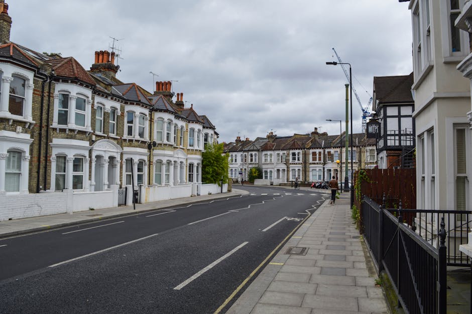 A quiet residential street with a row of Victorian-style terraced houses featuring bay windows, brick facades, and chimneys under a cloudy sky. The pavement runs alongside the houses, with a black metal fence visible in front of one property on the right. The street has a single lane of traffic, marked with dashed white lines, and a few parked cars are visible further down. A person wearing dark clothing is walking along the sidewalk, and a large construction crane is seen in the distance, indicating ongoing building work nearby. The scene captures a typical urban environment suitable for home relocation or furniture transport, with no visible furniture, boxes, or moving equipment in the image, but it illustrates the surroundings where a professional removals service, such as Catford Man and Van, could operate within this area.