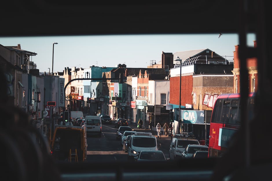 View of a busy urban street in Catford, with a mix of small retail shops and residential buildings lining the pavement. Several cars and a red double-decker bus are visible on the road, indicating transportation activity typical during a home relocation or moving day. The scene is captured from the back of a vehicle, with dark interior framing the image. The sky is clear, providing natural light that illuminates the facades of buildings made of brick and concrete, some with visible signage. Pedestrians are walking along the sidewalk, and a few are nearby waiting or carrying items, suggesting movement and logistics associated with furniture transport or packing and moving services. Occasionally, a van or delivery vehicle is seen parked close to the shops, reinforcing the context of local urban removals or household relocation in Catford. Catford Man and Van’s presence in the broader context can be implied through the scene’s setting focused on streets suitable for moving operations.