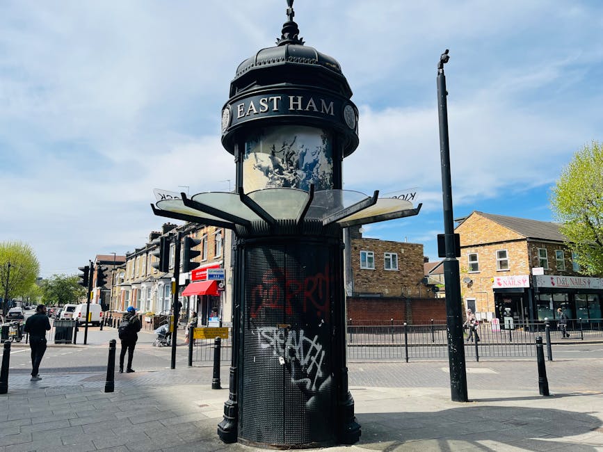 A historic black and silver cylindrical street clock with a domed top, situated on a sidewalk at a busy intersection in East Ham, London. The clock features a circular face with Roman numerals and decorative detailing, mounted on a sturdy metal column. Surrounding the clock is a cylindrical grill with graffiti tags, and the top of the structure displays the text 'EAST HAM.' In the background, there are several two-story brick and stone buildings housing shops, including a nail salon and a dental practice, with pedestrians walking along the pavement and crossing the street. The scene is illuminated by natural daylight, with a light blue sky featuring some clouds and a few green-leaved trees visible on the right side of the image. The area appears to be a lively urban commercial and transport hub, relevant to the local context of house removals and relocating services, as provided by Catford Man and Van.